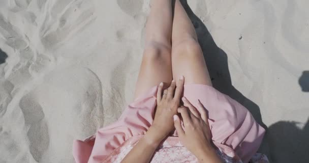 Mujer descansando en la playa de arena. El viento balancea la tela ligera de la falda rosa. La mujer sostiene la ropa con sus manos bronceadas. Disparos desde detrás del hombro . — Vídeos de Stock