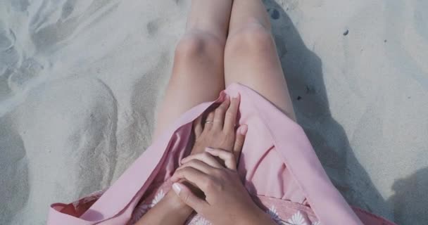 Donna che riposa sulla spiaggia di sabbia. Il vento ondeggia tessuto leggero di gonna rosa. La donna tiene i vestiti con le mani abbronzate. Sparare da dietro la spalla . — Video Stock