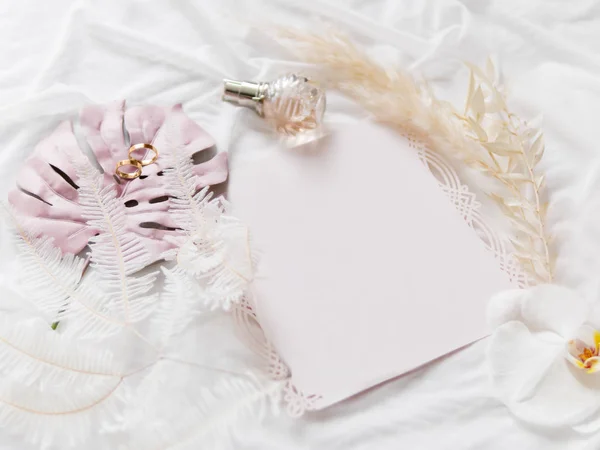 淡いピンクの明確な招待状と黄金の結婚指輪。ファラエノプシスオーキッド、コルタデリアセロアナ(パンパスグラス)と白い生地にピンクのモンテラの葉。折りたたまれた織物のコピースペースの招待と花を持つ優雅な背景. — ストック写真
