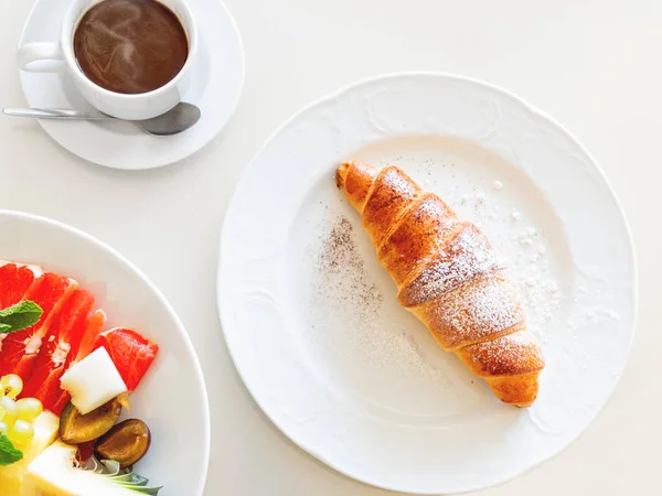 Continentaal ontbijt-smakelijke croissant, witte kop met warme koffie en bord met vers fruit. Top View. — Stockfoto