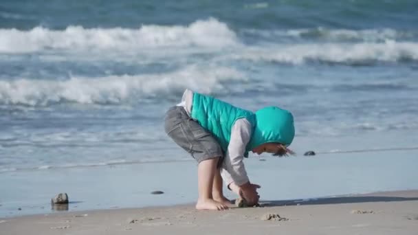 Mały chłopiec w kamizelce bawi się piaskiem od strony morza. — Wideo stockowe