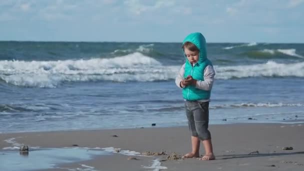 Batole chlapec ve vestě si hraje s pískem na mořské straně. — Stock video