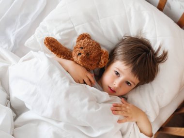 Bebek, tatlı oyuncak ayıyla yatakta yatıyor. Beyaz battaniyenin altındaki tüylü oyuncaklı küçük çocuk. Pelüş gardiyan çocuk uykusuna dikkat eder. Rahat evde sabah yatma vakti..