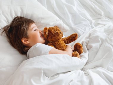 Bebek, tatlı oyuncak ayıyla yatakta yatıyor. Beyaz battaniyenin altındaki tüylü oyuncaklı küçük çocuk. Pelüş gardiyan çocuk uykusuna dikkat eder. Rahat evde sabah yatma vakti..