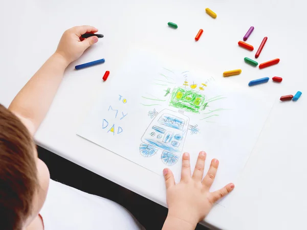 Left-handed toddler draws greeting card with funny robot for Father\'s day or Daddy\'s Birthday. Kid uses wax crayons. Top view on child\'s hands and pencils on white background.