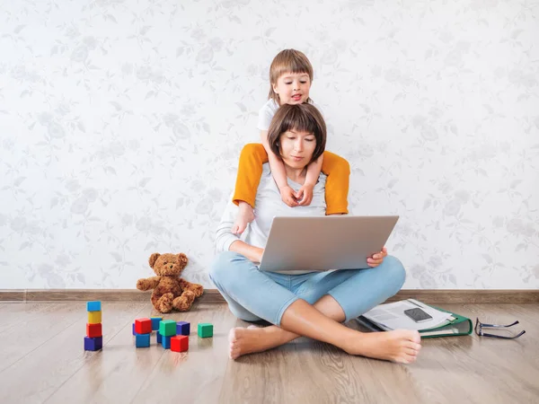 Mom and son at home. Mother works remotely with laptop, son sits on her neck. Freelance work in same time with raising children.