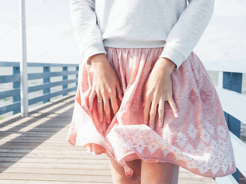 Woman stands on a pier by the sea and holds her skirt, which sways in the wind. Airy summer photo on seaside.