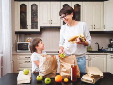 Kadın ve küçük çocuk mutfaktaki alışverişleri hallediyor. Çocuk elmayı ısırıyor. Kese kağıdında market teslimatı. Marketten abonelik hizmeti. Anne ve oğlu mutfakta..