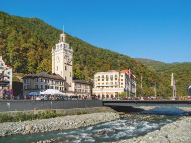 SOCHI, Rusya - 13 Ekim 2018. Turistler Rosa Dolina 'daki Rosa Meydanı' nda saat kulesinin yanında yürüyorlar. Soçi yakınlarındaki ünlü Rosa Khutor Alpine Kayak Merkezi.