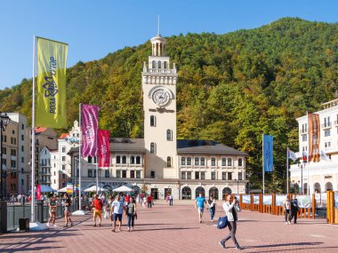 SOCHI, Rusya - 13 Ekim 2018. Rosa Dolina 'daki Rosa Meydanı' ndaki saat kulesinin yanında turistler yürüyor. Soçi yakınlarındaki ünlü Rosa Khutor Alpine Kayak Merkezi.