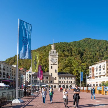 SOCHI, Rusya - 13 Ekim 2018. Rosa Dolina 'daki Rosa Meydanı' ndaki saat kulesinin yanında turistler yürüyor. Soçi yakınlarındaki ünlü Rosa Khutor Alpine Kayak Merkezi.