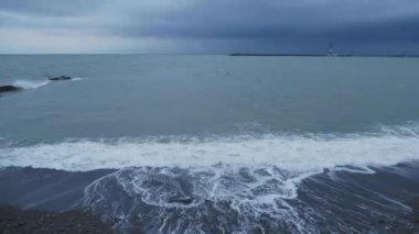 Martılar ve karabataklar dalgakıranların üzerinde otururlar. Fırtınalı arka planda deniz feneri. Soçi Limanı, Rusya.