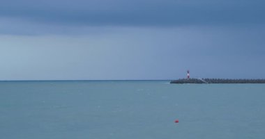 Martılar ve karabataklar dalgakıranların üzerinde otururlar. Fırtınalı arka planda deniz feneri. Soçi Limanı, Rusya.