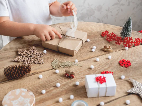 Child wrapped Christmas presents in craft paper with white fluffy pompons, feathers and fringe as decoration. Wooden table with hand made New Year gifts.