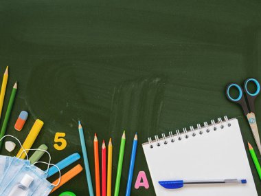 Renkli kırtasiye malzemeleri, koruyucu tıbbi maskeler ve el dezenfektanı. Okula dönüş konsepti. Kopya uzayı olan yatay karatahta arkaplan. Okul alışverişi. Yeni normal.