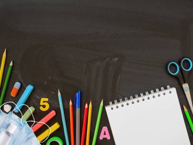 Renkli kırtasiye malzemeleri, koruyucu tıbbi maskeler ve el dezenfektanı. Okula dönüş konsepti. Kopya uzayı olan yatay karatahta arkaplan. Okul alışverişi. Yeni normal.