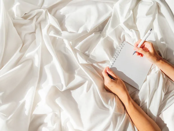 Woman is preparing to write or draw in diary. Top view on crumpled bed sheet and notepad with white paper pages. Practice of positive writing at morning after waking up. Flat lay with copy space. — Stock Image Woman Preparing Write Draw Diary Top View Crumpled Bed Sheet Stock Picture