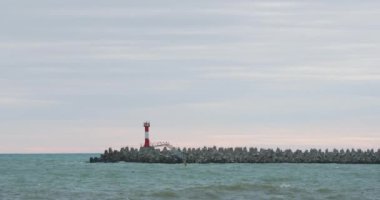Martılar ve karabataklar dalgakıranların üzerinde otururlar. Günbatımı arka planında deniz feneri. Soçi Limanı, Rusya.