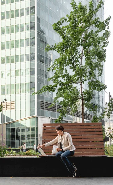 Business woman with short haircut and eyeglasses sits on bench in urban park with laptop and cardboard cup of coffee. Student learns remotely from outdoors. Modern Internet technologies. Video call.