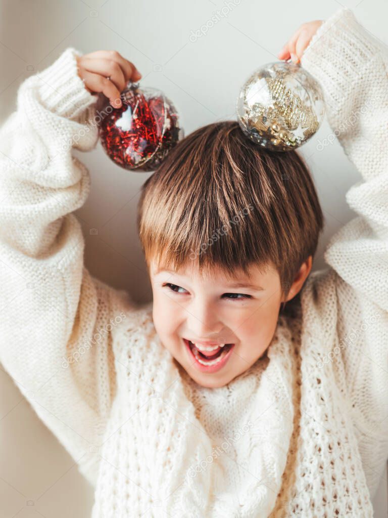 Kid with decorative balls for Christmas tree.Boy in cable-knit oversized sweater.Cozy outfit for snuggle weather.Transparent balls with red, golden spangles inside.Winter holiday spirit.New year.