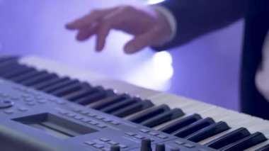 Takım elbiseli bir adam konserde altı telli elektrikli sentezleyici çalar. Müzisyenlerin ellerindeki flaş disko ışıklarıyla yakın çekim görüntüleri..