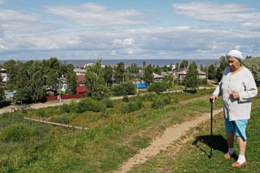 Belozersk, Rusya Federasyonu - 05 Ağustos 2015: Yaşlı bir kadın ile bir baston köy ve Belozersk gölde arka plan üzerinde duruyor