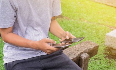 Park'ta her iki elinde iki smartphone kullanarak ahşap sandalye üzerinde oturan adam