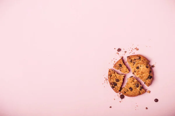 Broken chocolate chip cookie and crumbs on pink background.