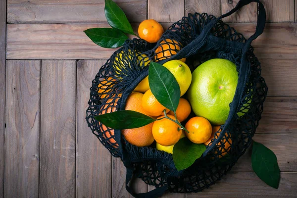 Multicolored citrus fruits in textile bag on old wooden table. Healthy food and zero waste concept.