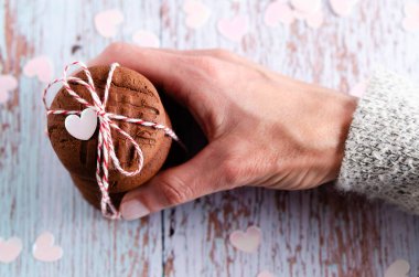 Bir kadın eli, kırmızı ve beyaz bir ip ve ahşap açık mavi arka planda küçük pembe kalplerle bağlanmış yığınla çikolatalı kurabiyeyi tutuyor. Üst görünüm.