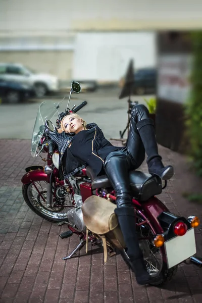 woman with blond hair in black high boots, leather jacket lying on old, vintage, retro motorcycle