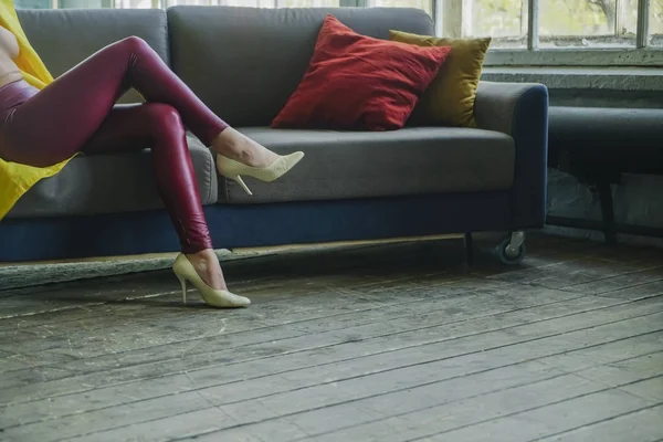 赤い革のズボンと白い低い靴を身に着けている女性のディテール 灰色のコーチは Pilliows が付いているソファーの上に女性座る — ストック写真