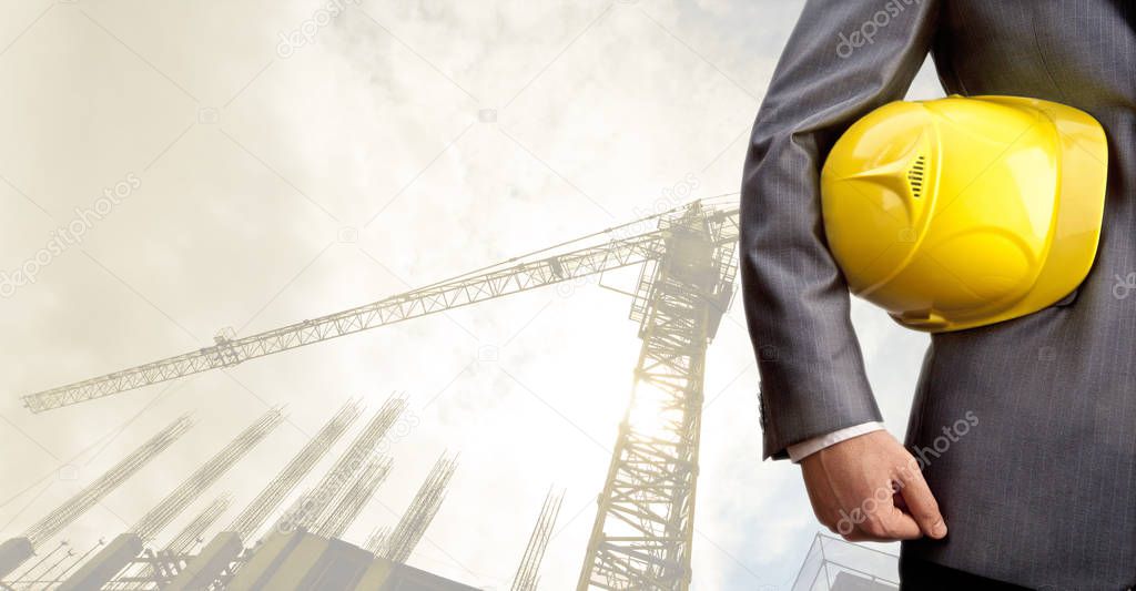 engineer holding yellow helmet for workers security on background of new  building and construction cranes on background of evening sunset cloudy sky