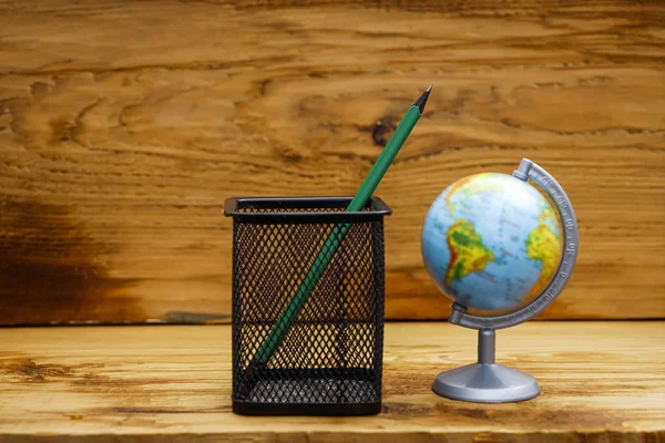one green pencil in metal net case and globe against wooden background. back to school