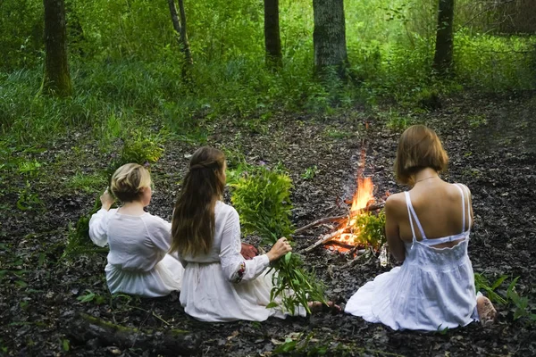 Focus on flame. three witches sitting near fire in evenig forest and  holding in hands wild spring flowers.