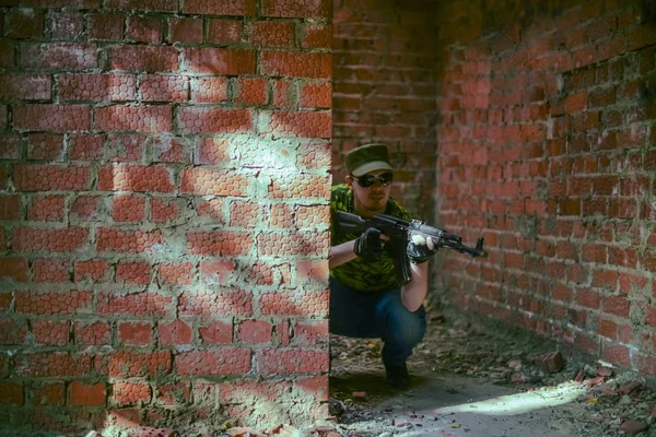 focus on wall. Caucasian military man in  black sunglasses indoor urban room space stand with machine gun near abandoned red brick wall. Dusk light. Corridor in perspective.Empty space for inscription