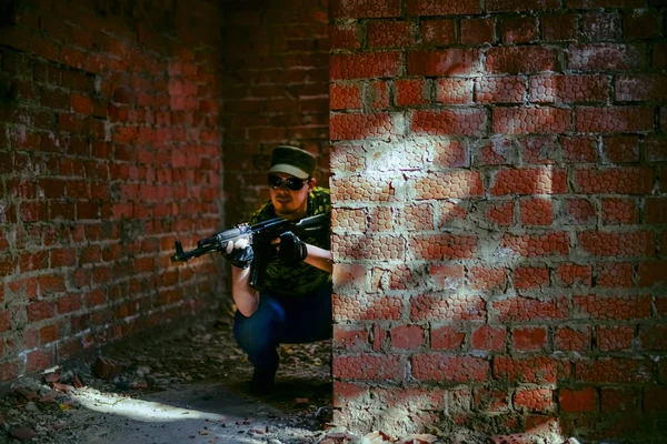 focus on wall. Caucasian military man in  black sunglasses indoor urban room space stand with machine gun near abandoned red brick wall. Dusk light. Corridor in perspective.Empty space for inscription
