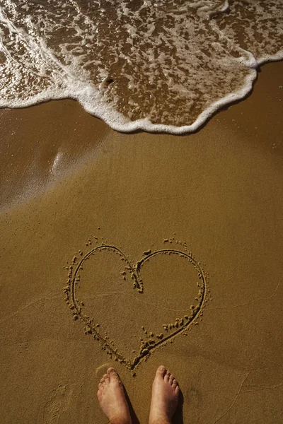 Young man Drawing A Heart In The Sand With  Foot. Male feet and love heart shape on a yellow sandy beach.
