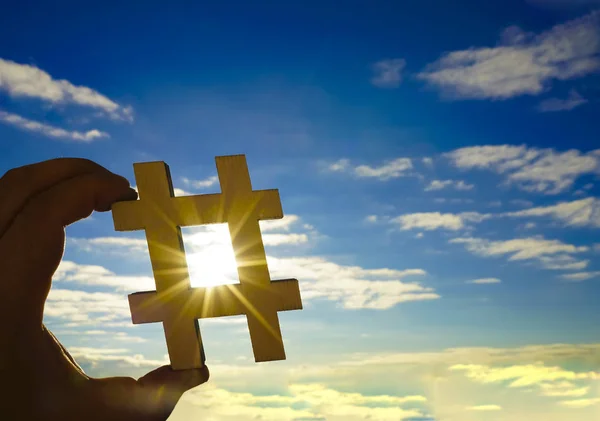 closeup of the hand of a young caucasian man  holding a wooden sign in the shape of a hash tag symbol against the sky, with some blank space. hashtag