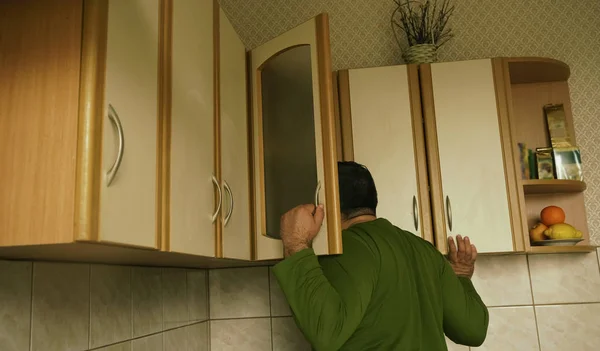Young man in sweater peeking  into kitchen cupboard at home, viewed from the back side. alone hungry man search food.