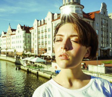 Portre büyüleyici kısa saçlı esmer kız beyaz bir t-shirt, gözleri kapalı, parlak bir Güneşli günde bir nehir tarafından beton bir iskelede güneş ışığı zevk