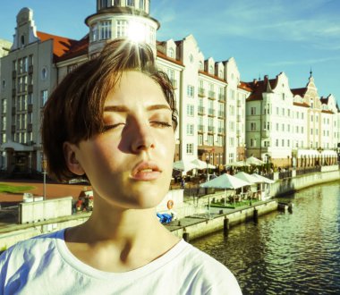 Portre büyüleyici kısa saçlı esmer kız beyaz bir t-shirt, gözleri kapalı, parlak bir Güneşli günde bir nehir tarafından beton bir iskelede güneş ışığı zevk