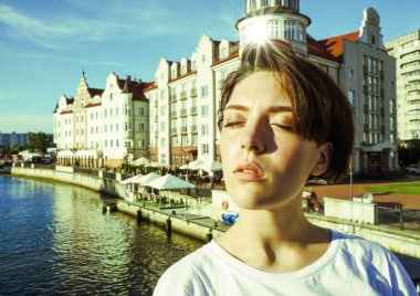 Portre büyüleyici kısa saçlı esmer kız beyaz bir t-shirt, gözleri kapalı, parlak bir Güneşli günde bir nehir tarafından beton bir iskelede güneş ışığı zevk