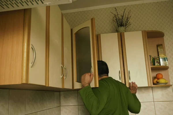 Young man in sweater peeking into kitchen cupboard at home, viewed from the back side. alone hungry man search food.