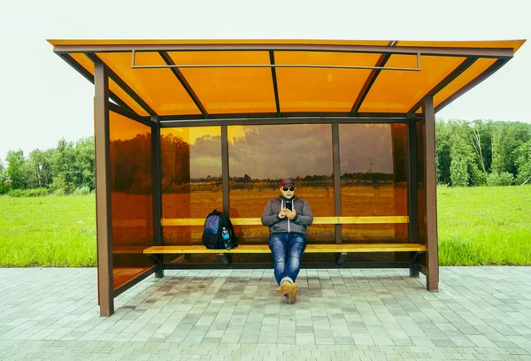 Радісний молодий чоловік чекає на транспорт на зупинці. чоловік сидить — стокове фото