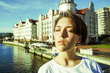 Portre büyüleyici kısa saçlı esmer kız beyaz bir t-shirt, gözleri kapalı, parlak bir Güneşli günde bir nehir tarafından beton bir iskelede güneş ışığı zevk