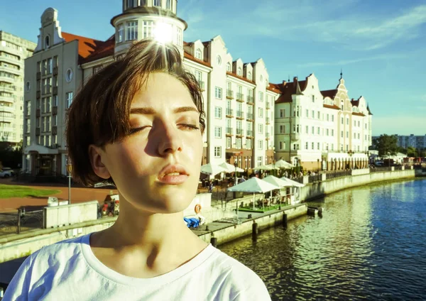 Portre büyüleyici kısa saçlı esmer kız beyaz bir t-shirt, gözleri kapalı, parlak bir Güneşli günde bir nehir tarafından beton bir iskelede güneş ışığı zevk