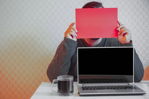 Young man closed face with blank red banner. Man holding advertising sheet, copy space. Business man hiding behind a white banner on a gray background. empty screen of laptop.
