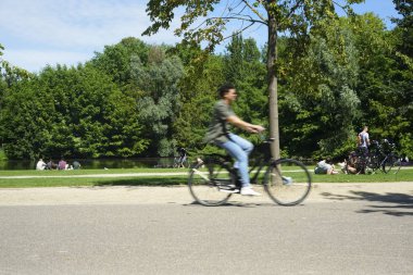 Sıcak bir yaz gününde Vondelpark. İnsanlar güneşin altında ağaçların altında çimenlerin üzerinde dinleniyorlar. Bisikletler yola çıkıyor. Amsterdam, North-Holland, Hollanda 