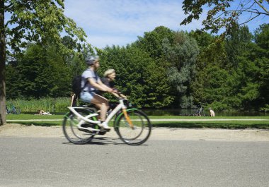 Vondelpark sıcak bir yaz gününde. İnsanlar güneşin altında ağaçların altında çimenlerin üzerinde dinleniyorlar. yolda iki Bisiklet sürme çift. Amsterdam, North-Holland, Hollanda 